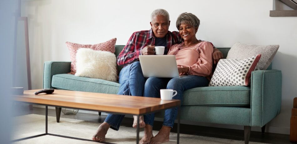 Older couple sitting on a green couch having coffee and smiling at a laptop.