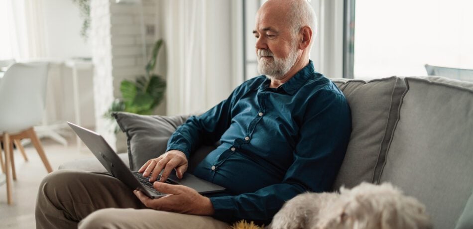 Older man wearing a blue shirt holding an open laptop with his dog next to him on the couch.