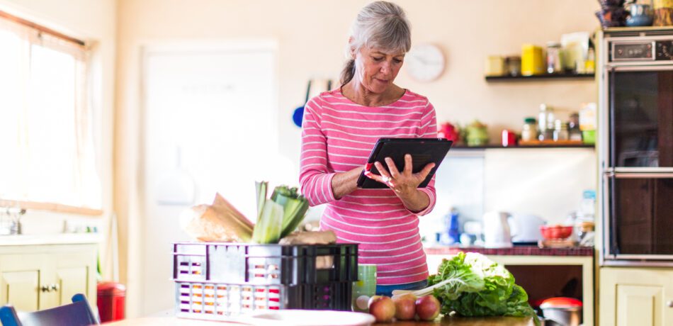 An older woman in a pink top, looking at an iPad while groceries sit on the table in front of her.