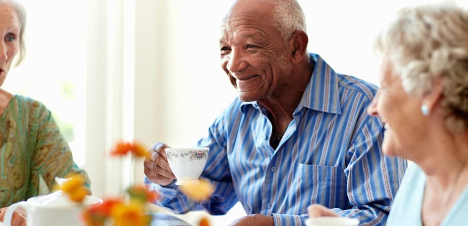 An older man wearing a blue striped shirt with two women either side of him as they all have tea together.