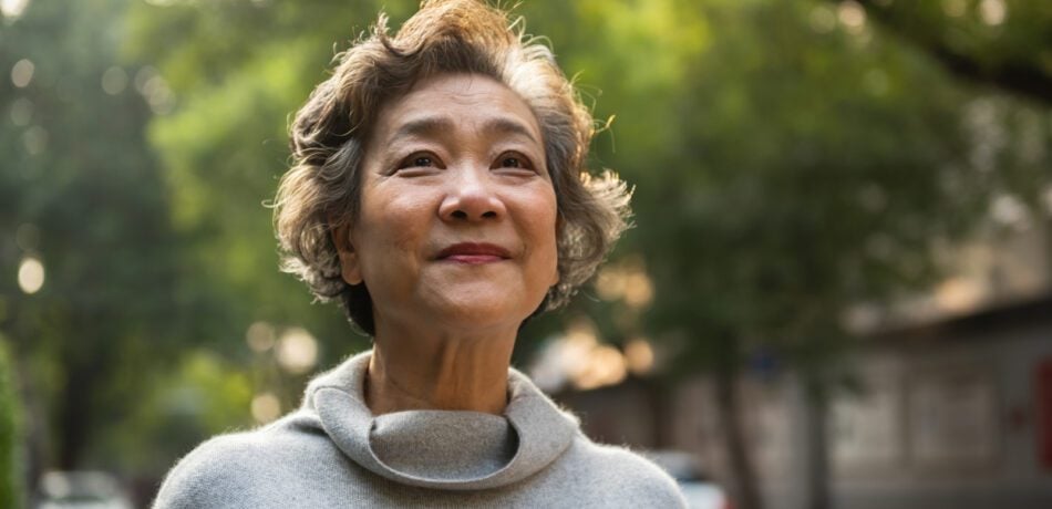 An older woman wearing a gray turtleneck top smiling while looking into the distance with trees behind her.