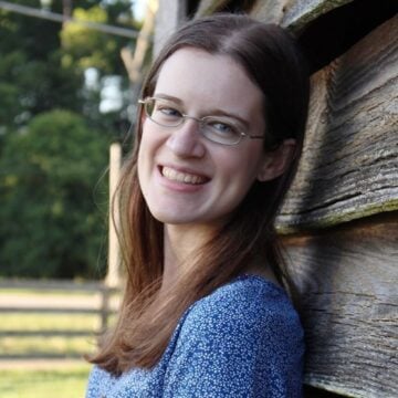 Headshot of woman in blue top and eyeglasses smiling at camera while leaning against wooden shed.