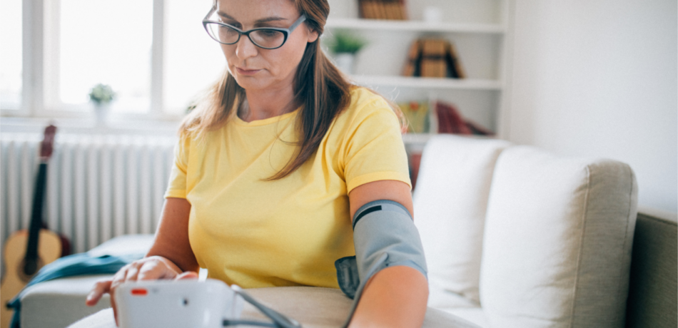 Woman in yellow t-shirt wearing glasses is sitting at table using a blood pressure cuff to take her own readings at home.