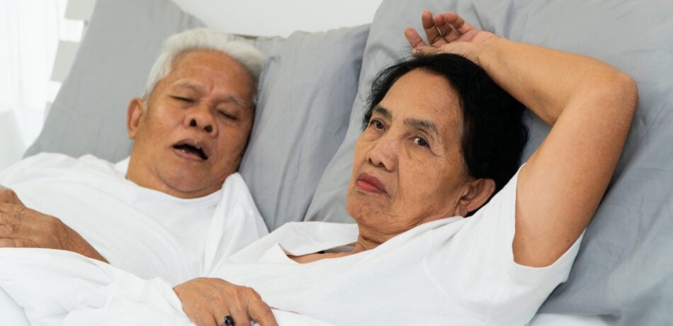 Older woman laying awake in bed next to her husband who is snoring.