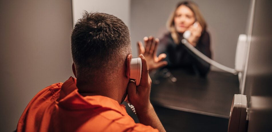 A male prisoner on a phone with his female visitor also on a phone as they touch the glass between them with their hands.