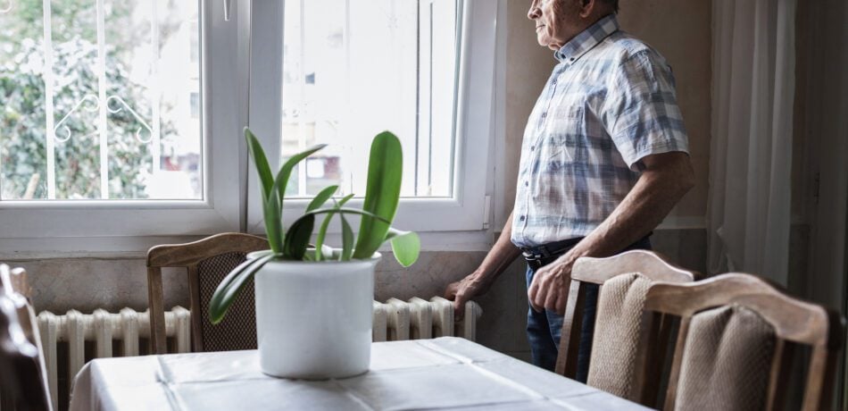 Older man standing next to a table with a plant on it while he looks out of his window with a somber look on his face.