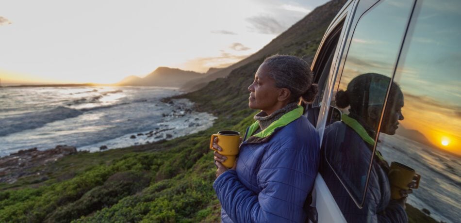 Mature woman enjoying sunset by the sea while leaning against her vehicle holding a travel mug.