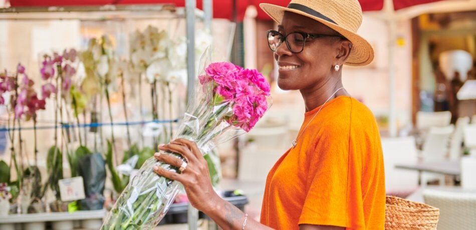 Woman wearing a hat and glasses smiling and holding a bunch of pink flowers in a farmer's market.