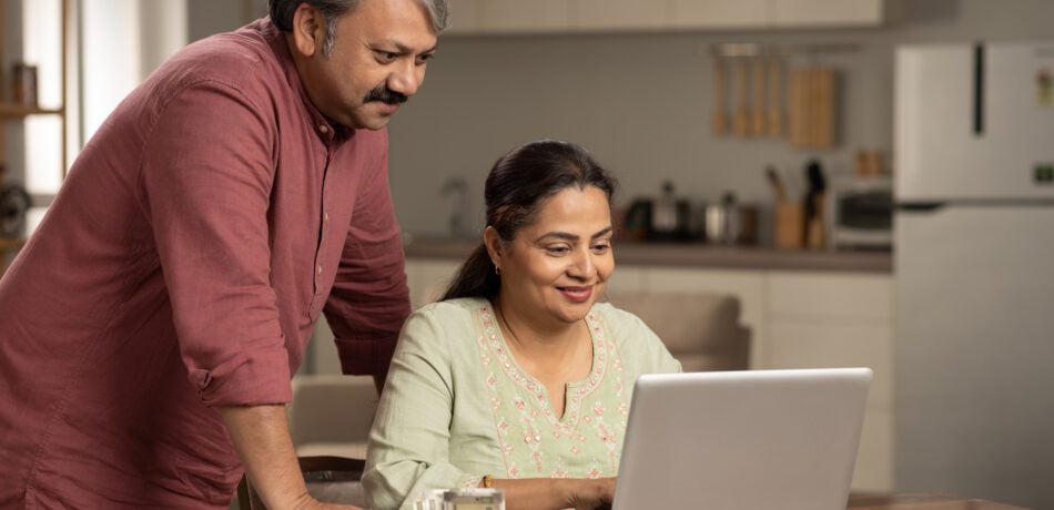 Indian couple smiling looking at laptop at kitchen table.