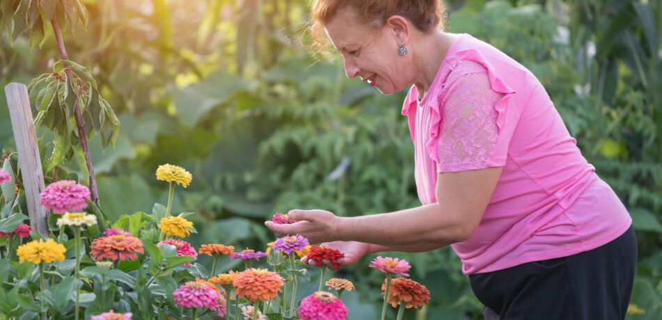 Older woman in pink shirt smiling while looking at her flower garden.