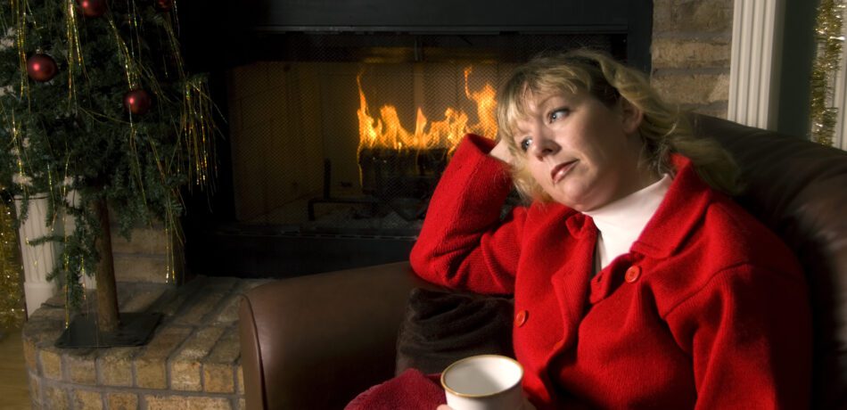 Woman in red jacket holding a mug sitting in front of a fireplace and small Christmas tree looking sad.