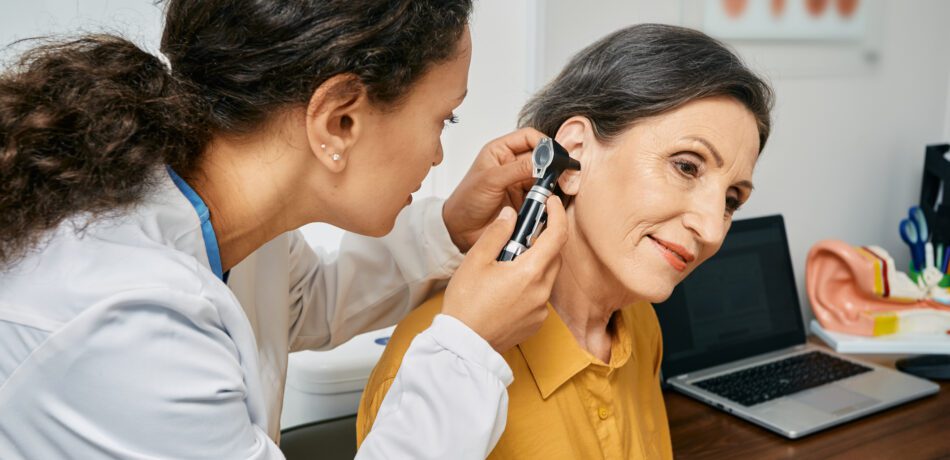 A female doctor using an otoscope to look in the ear of an older woman wearing a gold colored shirt.