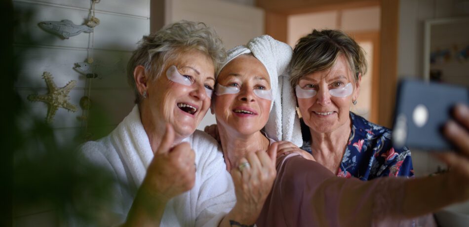 Three senior women friends in bathrobes taking a selfie while wearing undereye treatment patches for a spa day at home.