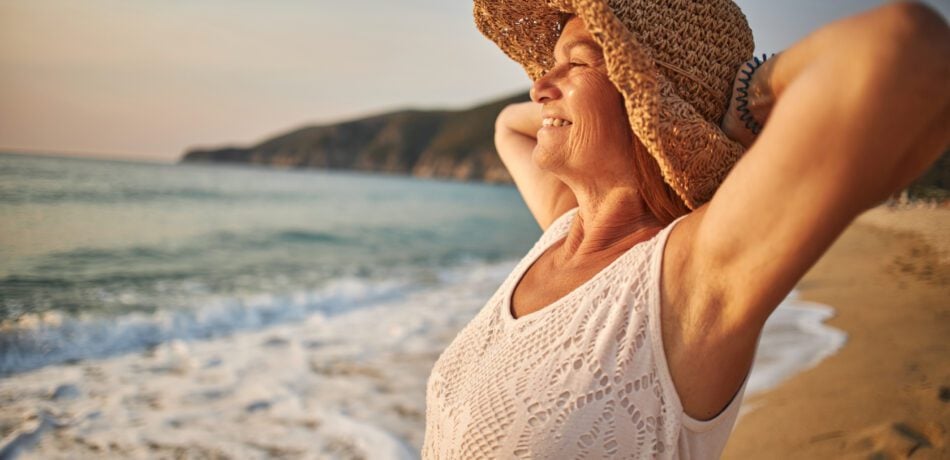 Senior woman wearing a large sunhat smiling standing on the beach while looking at the ocean.