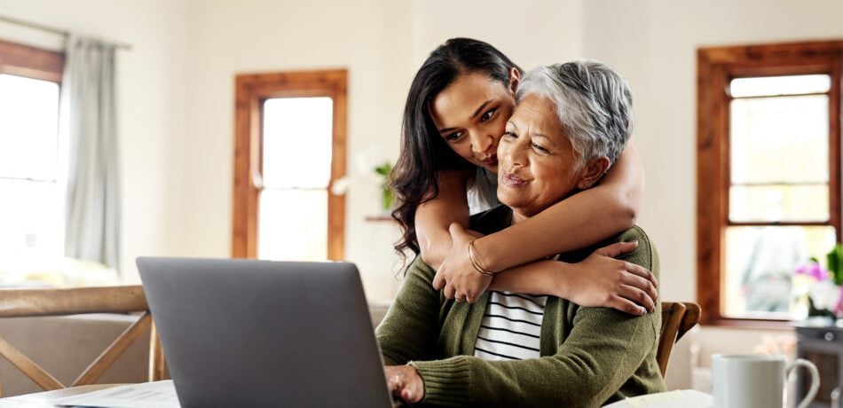 Young woman hugging an older woman as they smile and look at a laptop on the table.