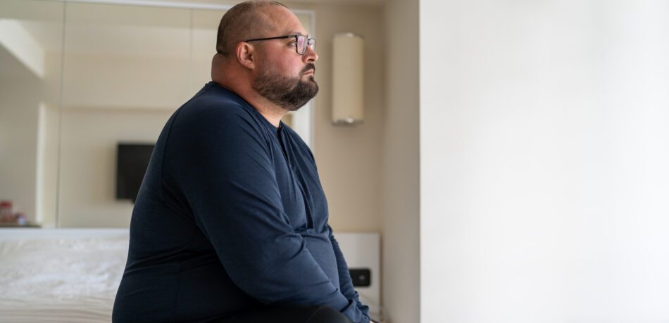 Lonely overweight man wearing glasses and a navy top sits on a bed looking sad.