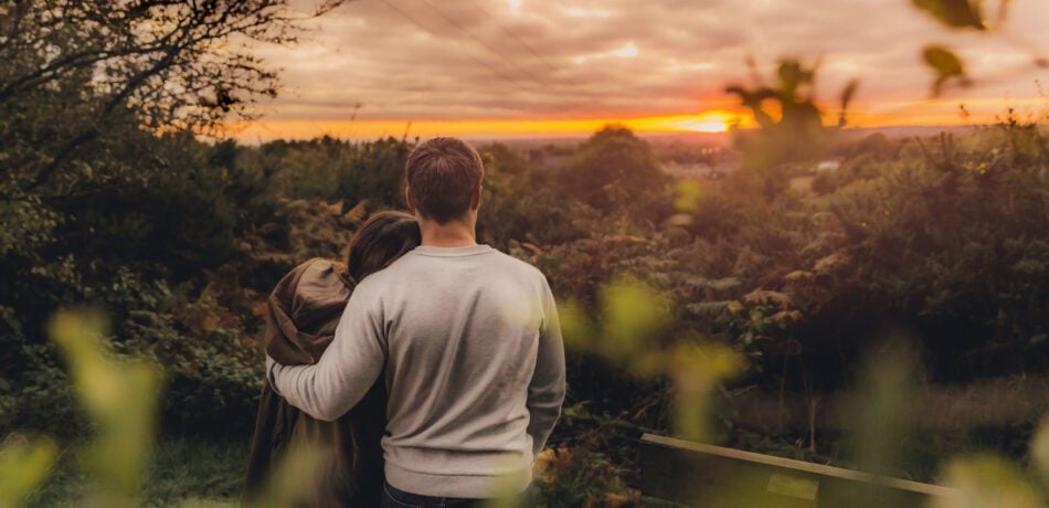 Back view of couple standing on a hill watching a sunset while the man has his arm around the woman's back.