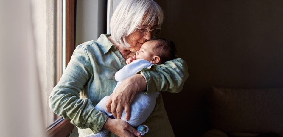 Grandmother holding and kissing her infant grandson by a window.