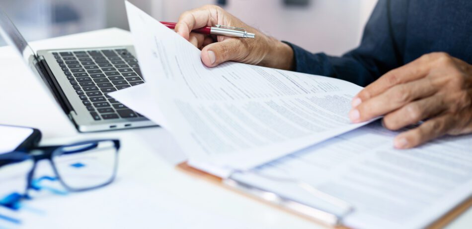 Man's hands on paperwork with open laptop and pair of eyeglasses on the desk.