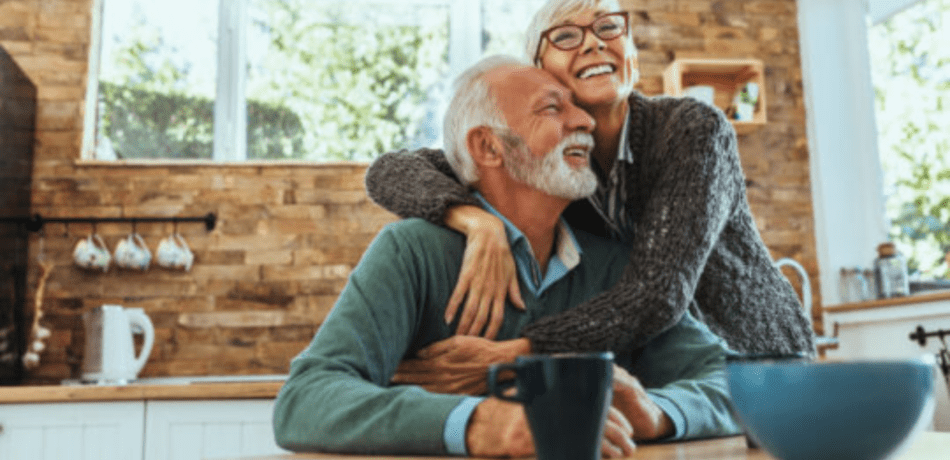 Older couple hugging and smiling while sitting at their kitchen table.