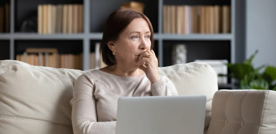 Woman looking pensive with her hand to her mouth as she sits looking into the distance while sitting on her couch with a laptop.
