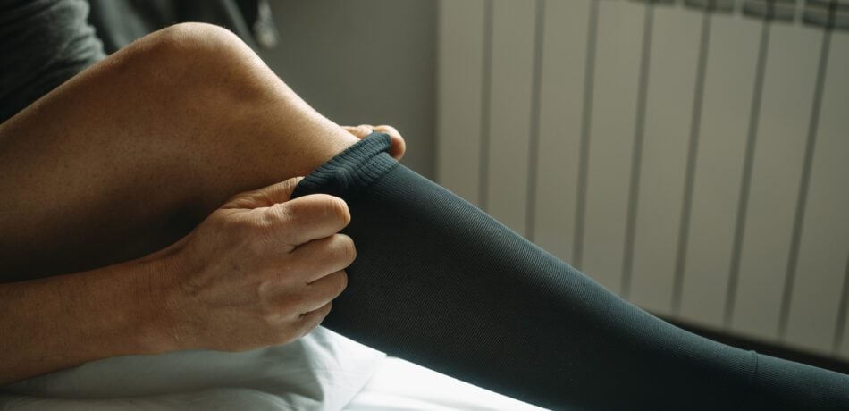 Man putting on a leg compression sock sitting on his bed at home.