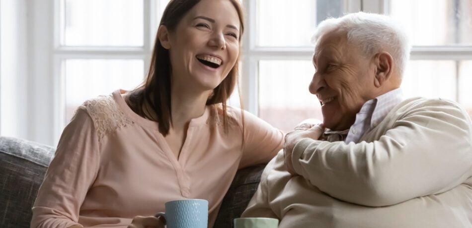 Adult daughter laughing having coffee on couch with her senior father.