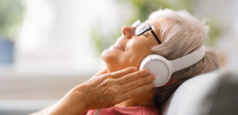 Senior woman listening to music in headphones sitting on the sofa in the room at home.