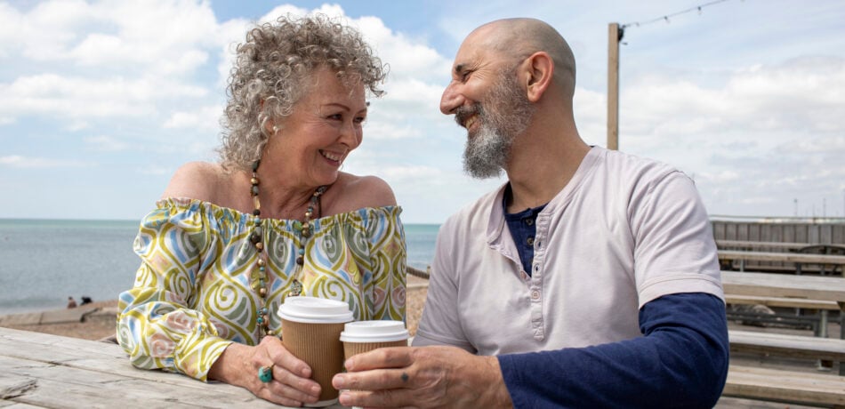 Mature couple enjoying coffee together by the beach at a picnic table.