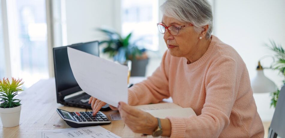 Older woman looking serious as she holds a paper and uses a calculator in front of a laptop on her desk.