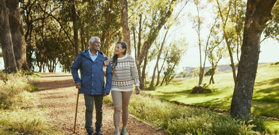 An elderly couple smiles and walks arm in arm through a park, surrounded by lush trees and bathed in warm sunlight.