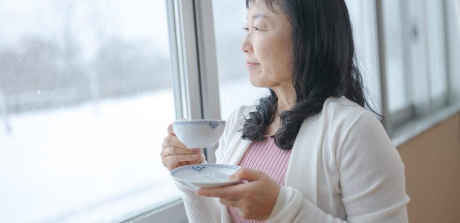 Older Asian woman looks out of her window to the snowy yard while holding a teacup and enjoying a cup of tea.