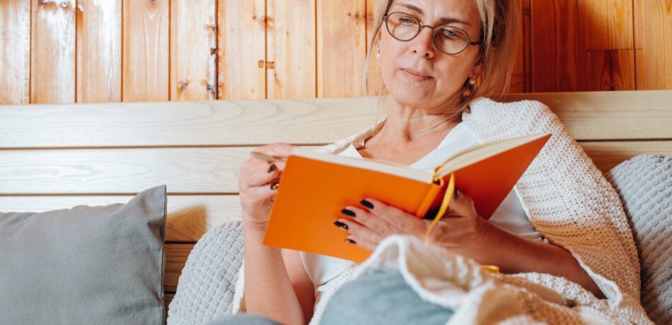 Middle aged woman wearing glasses taking notes in an orange notebook while cozied up on a couch with a throw blanket over her.