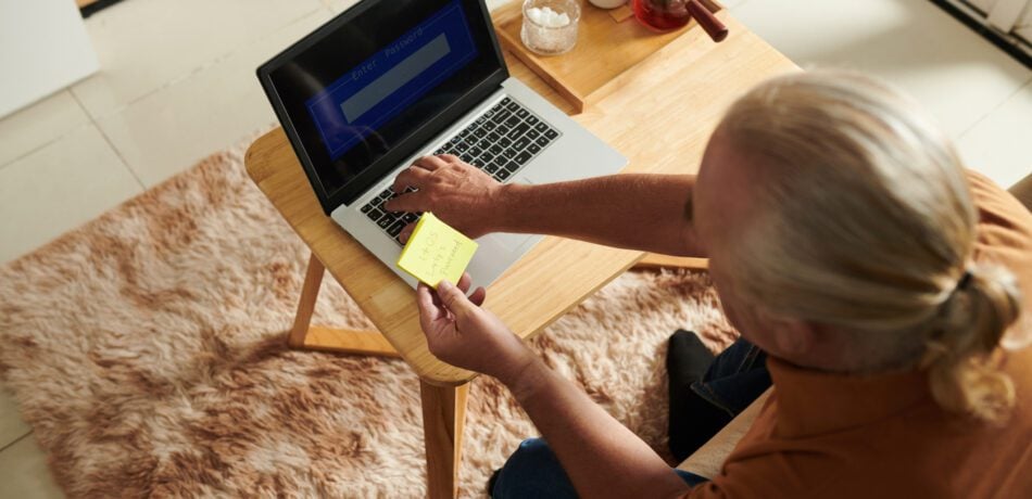 Senior man entering password written on sticky note into his laptop.