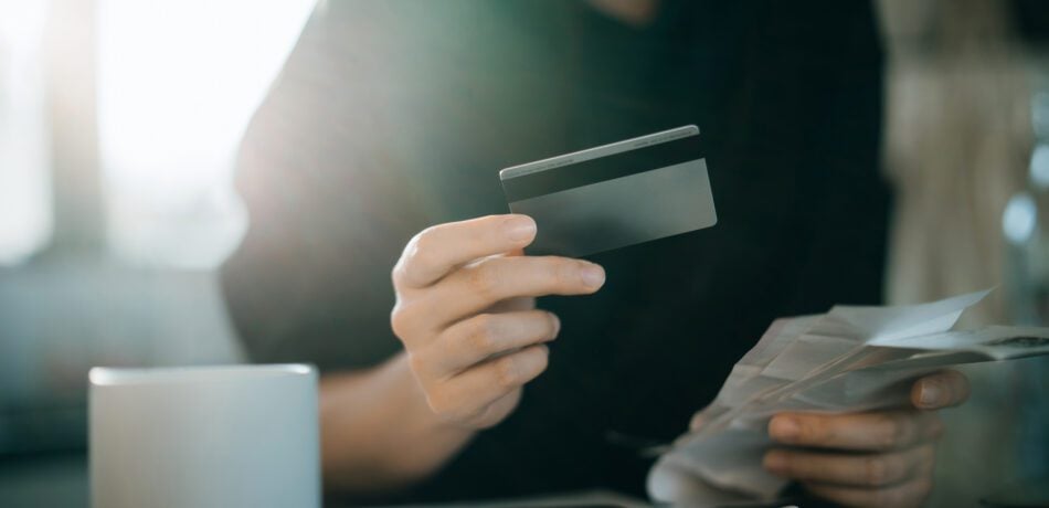 Cropped shot of woman's hand holding credit card and going over bills with a mug of coffee next to her.