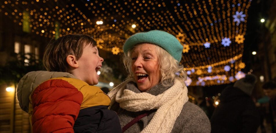 Grandmother holding her young grandson under festive holiday lights at night. Both are outside smiling and laughing together bundled up in cold weather jackets.