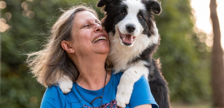 Border Collie dog on woman's shoulders posing with green trees in the background.