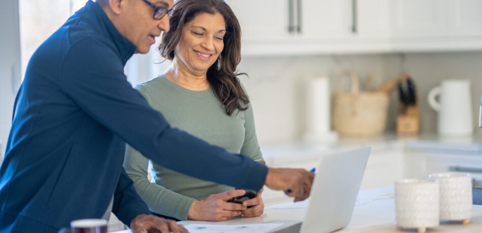 A middle aged couple stand at their kitchen island with an open laptop and paperwork smiling and working together on finances.