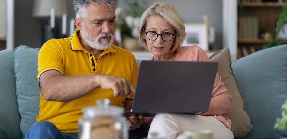 A retirement-aged couple is using a laptop, sitting together on a sofa in a living room.