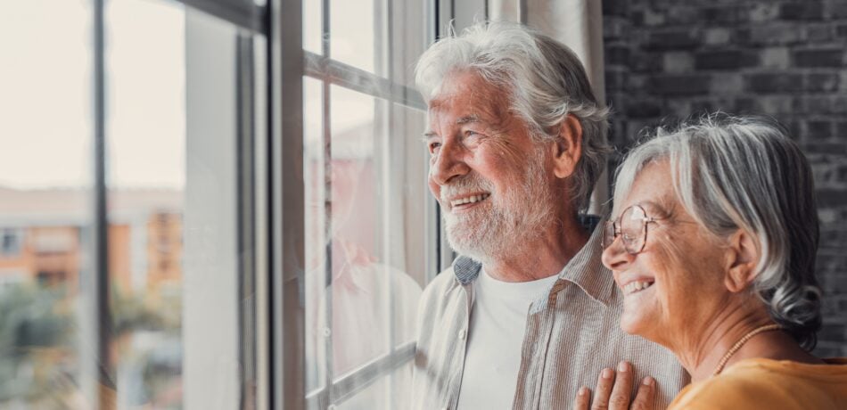 A senior couple happily looks out the window at home.